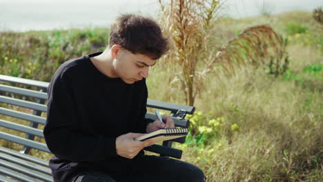 college student writing notes and studying on a bench near the sea
