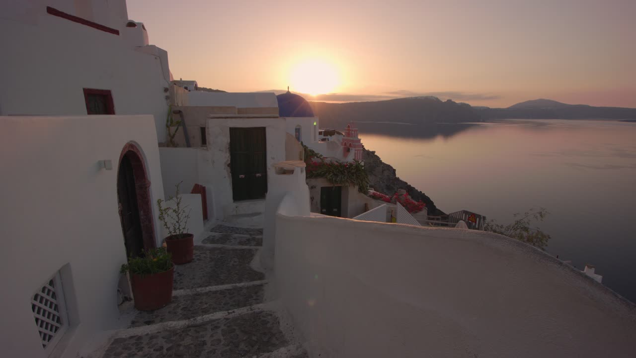 Golden sunset light bathes the whitewashed cliffside homes of Santorini, Greece. A peaceful stone alley winds through traditional Cycladic architecture, overlooking the calm Aegean Sea
