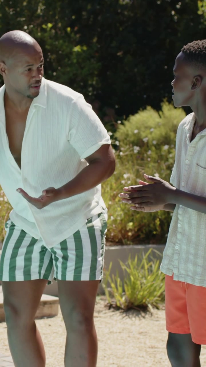 Video verticale: Adulto che cammina esortando un giovane con pantaloncini arancioni a estendere i palmi su un sentiero soleggiato, vicino al marciapiede