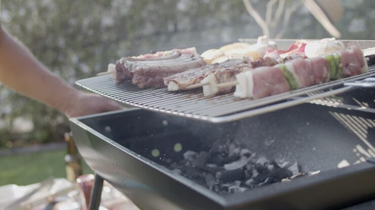 Pemanggang barbeku dengan daging dan pria mengaduk serta meniup bara arang.