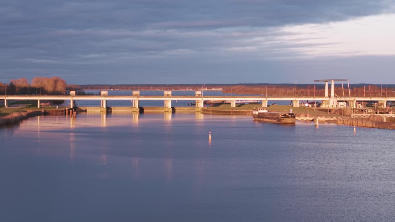 Aerial view of a river lock with a cargo ship, a modern bridge, and golden-hour reflections on the water.