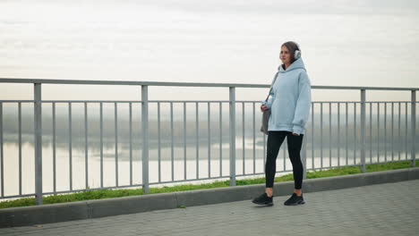 fair woman in casual wear walking slowly while listening to music with headphones, holding a bag, with water and green grass in the background, creating a serene and relaxed atmosphere