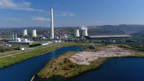 Thermal-power-plant-with-smoking-chimney-behind-the-lake-and-old-excavator,-bridge-on-the-right,-sunny-day