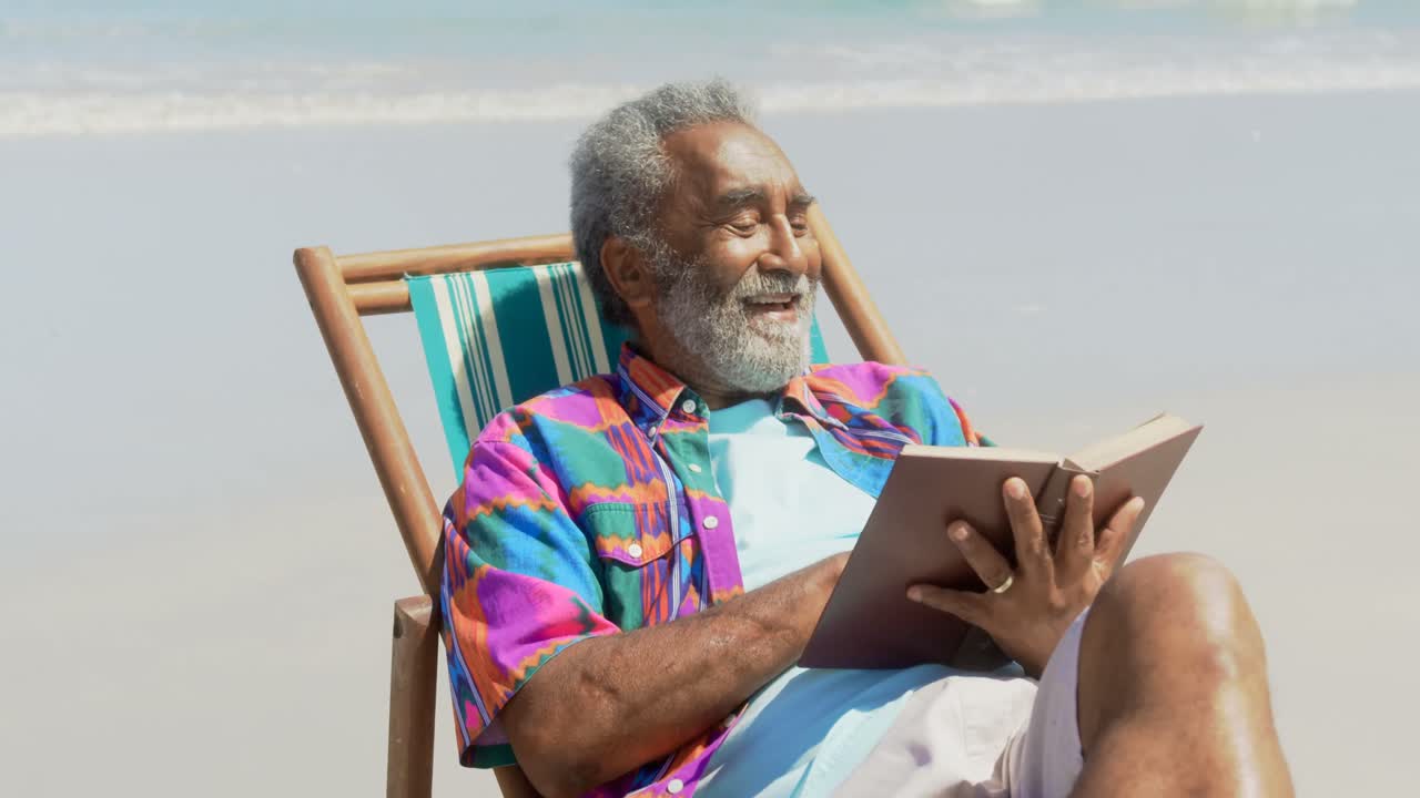vista frontal de un hombre afroamericano mayor activo leyendo un libro en una silla de sol en la playa 4k