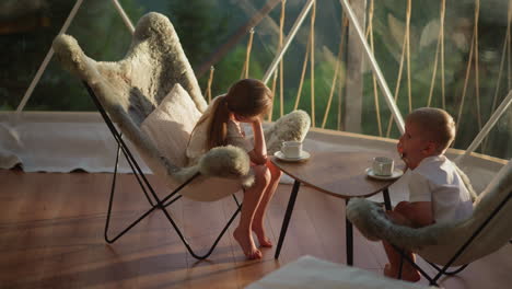 children rest sitting at table. brother and sister relax after breakfast looking at lighted window in comfortable glamping tent. family holiday