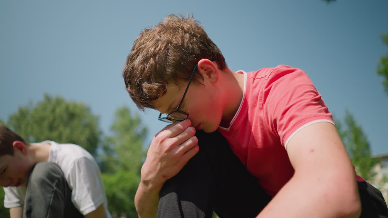 A young boy in a red shirt kneels down, adjusting his glasses as he focuses on a task outdoors, with a blur view of his brother kneeling close by, focused on what he is doing