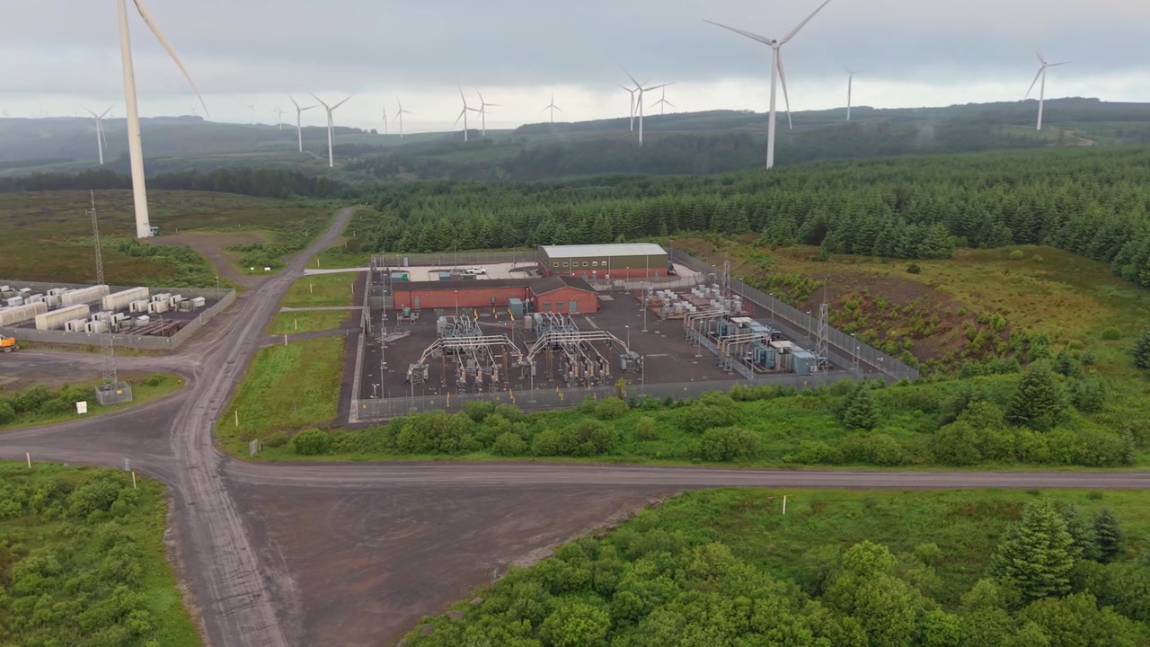 Wide View of Electrical Substation Managing and Re-Directing Flow of Electricity on Natural Landscape with Wind Turbines in Background.
