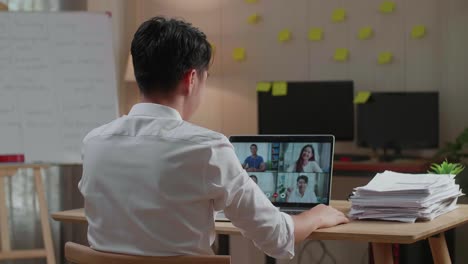 vista trasera de un hombre asiático haciendo una llamada de video en una computadora portátil mientras trabaja con documentos en la oficina