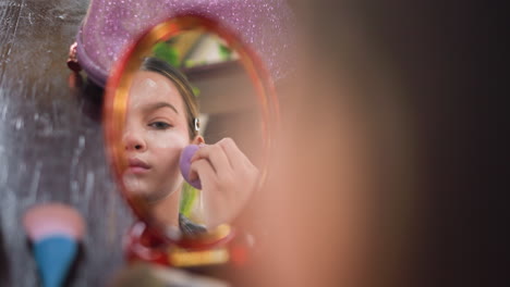 close-up mirror reflection of young girl applying powder to face with makeup sponge, blurred background with beauty tools and products on table showcasing makeup routine