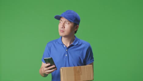 primer plano de un mensajero masculino asiático en uniforme azul caminando y usando un teléfono inteligente mientras entrega una caja de cartón en el fondo de la pantalla verde en el estudio