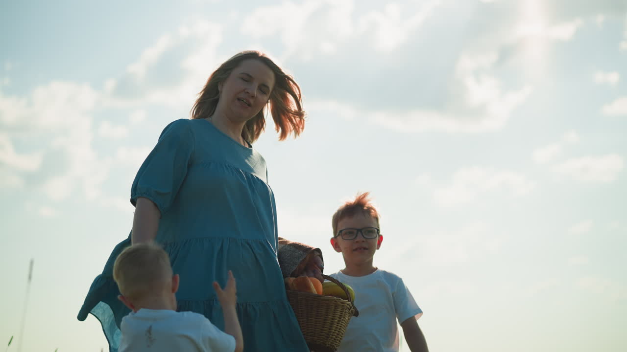 A joyful woman in a flowing blue gown walks through a green grass field with her two young sons, both in white shirts. She carries a basket while the children excitedly point at something in the grass