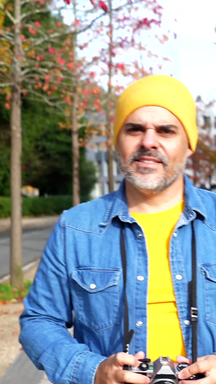 Man in denim jacket and yellow beanie holding a camera outdoors in autumn