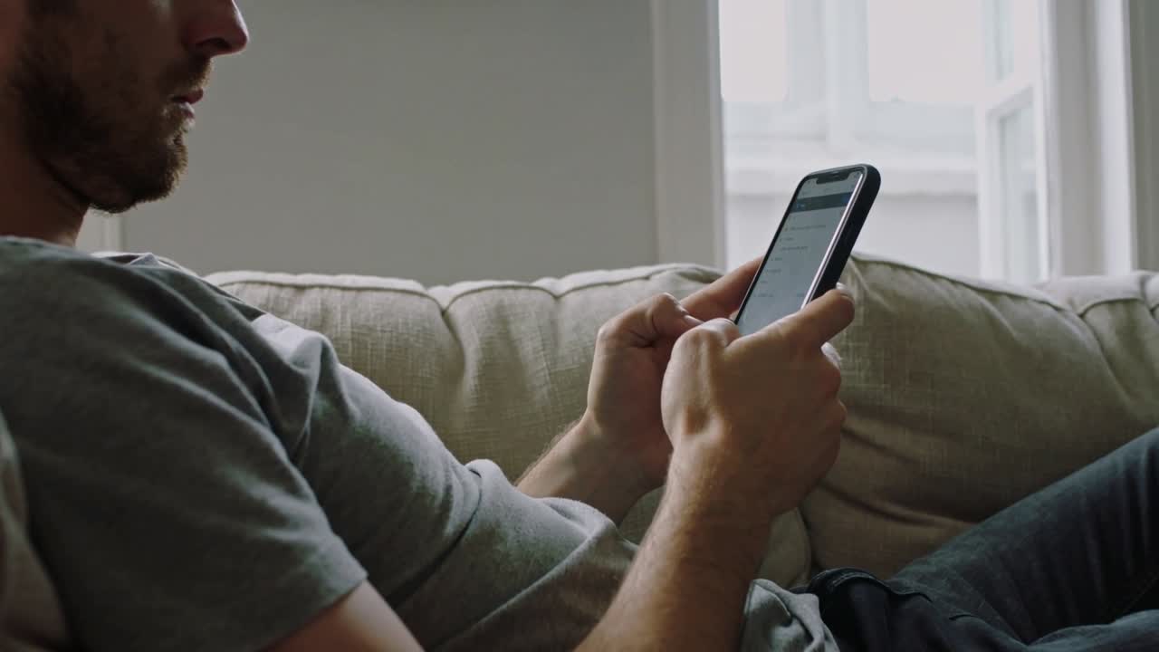 Muž opírající se dozadu klepe na chytrý telefon, kontroluje zprávy na béžové pohovce u okna, oblečený v šedém tričku.