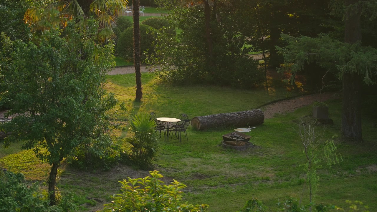 Vista estática de una acogedora zona de asientos de jardín con una mesa y sillas de acero, rodeada de árboles frondosos y suave luz natural dorada, creando un tranquilo refugio al aire libre.