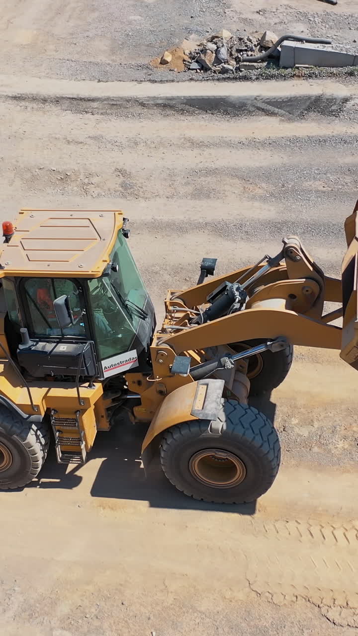 Excavator on the territory of asphalt manufacture outdoors. Modern excavator pouring out sand from bucket into a special tank. Top view. Vertical video