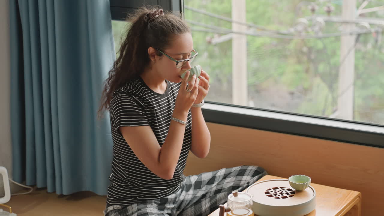 In una suite d'hotel, un ospite si gode uno spuntino e il tè vicino alla finestra. Un piatto di biscotti e una tazza in ceramica sono poggiati su un tavolino basso, con sedute informali a terra. All'esterno, un paesaggio verdeggiante e un'atmosfera pomeridiana rilassata.