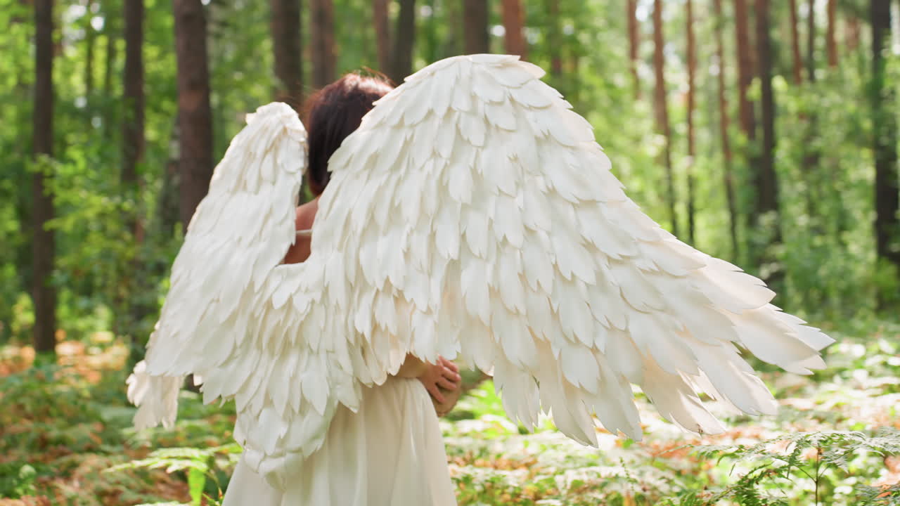 Back view of fairy goddess with large white angel wings walking gracefully through green forest, sunlight glowing over feathers as she moves peacefully