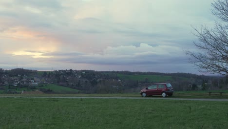 vista estática de mano del coche estacionado fuera del lado de la carretera del país con vistas a la aldea al atardecer