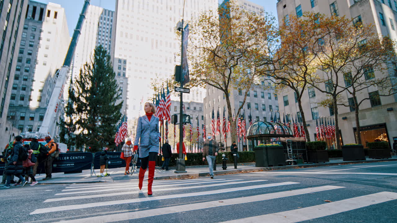 New York, USA, 5 October 2025: Wide shot of woman crossing street in Rockefeller Center. Woman in plaid suit and red boots crossing the street at Rockefeller Center with flags and trees