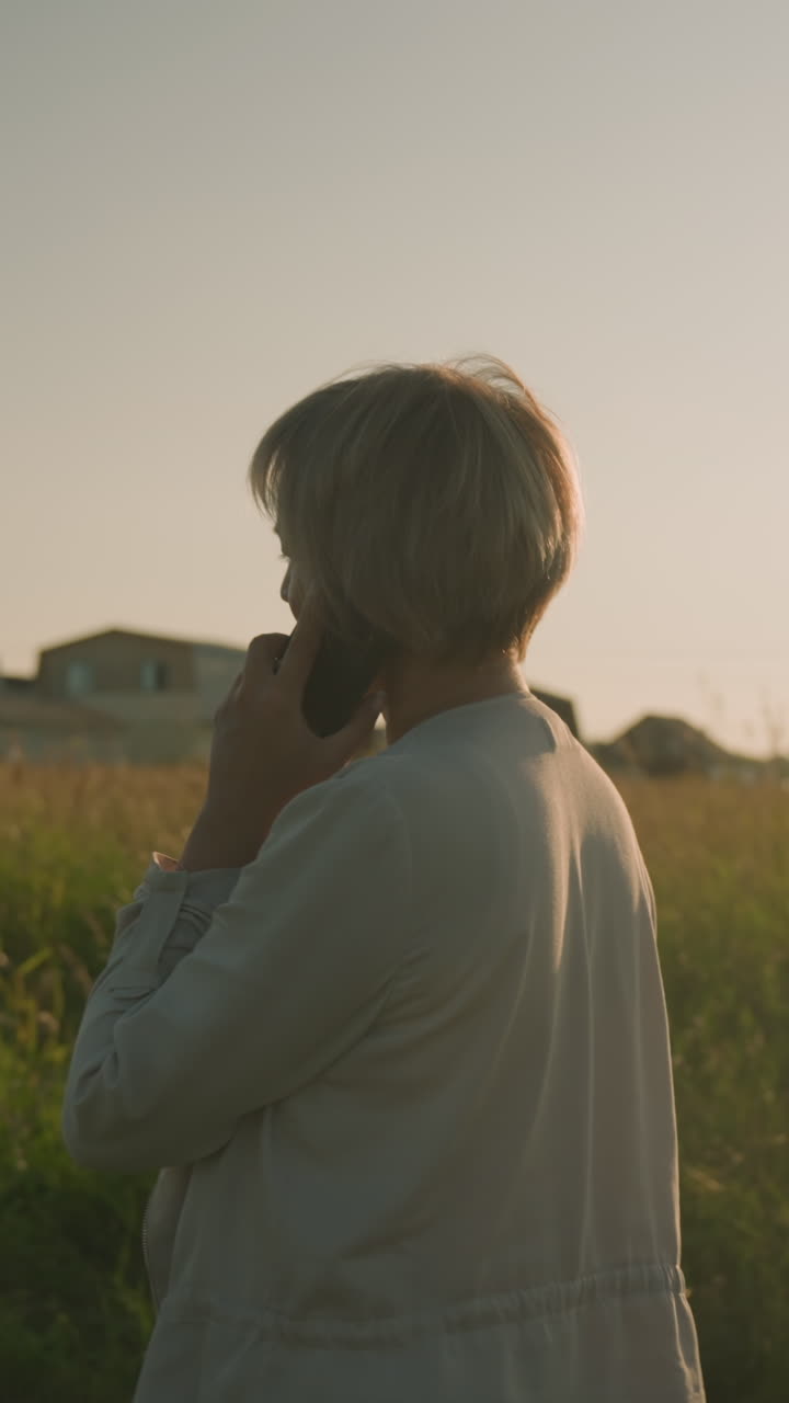 vista lateral de una mujer hablando por teléfono mientras camina cerca de un campo de hierba durante la hora dorada, edificios distantes visibles en el fondo, la luz del sol proyectando un brillo cálido sobre el paisaje rural