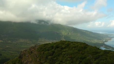 Toma-Aérea-De-La-Isla-Mauricio,-Con-Una-Montaña-En-Primer-Plano,-Una-Más-Al-Fondo,-Que-Desciende-Al-Mar