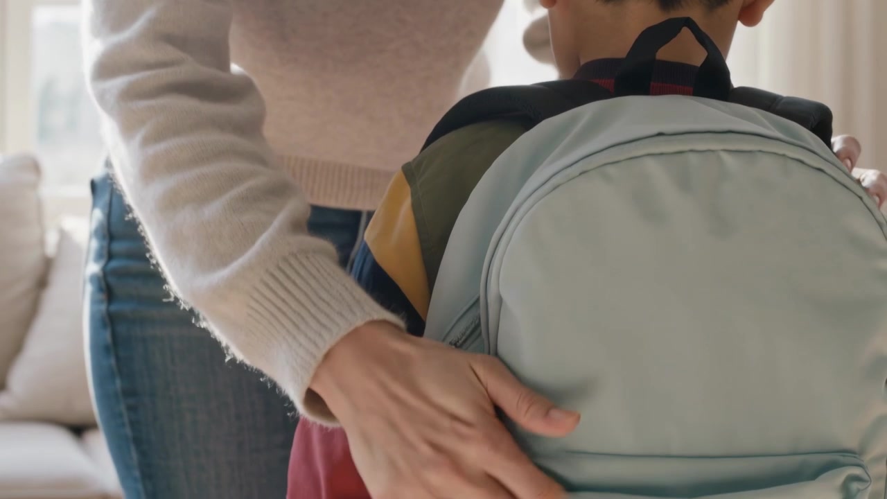 Adjusting mother in sweater securing light-blue backpack on child in red jacket at home for school