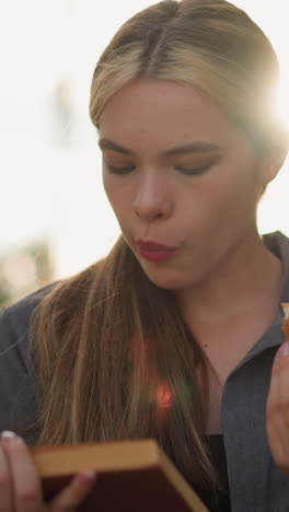 dama de camiseta gris leyendo un libro mientras sostiene una hamburguesa, masticando pensativamente con la suave luz del sol iluminando la escena, el fondo borroso por los rayos del sol y la vegetación