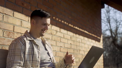 close-up view of caucasian man having a video call on laptop while sitting on a bench outside a country house