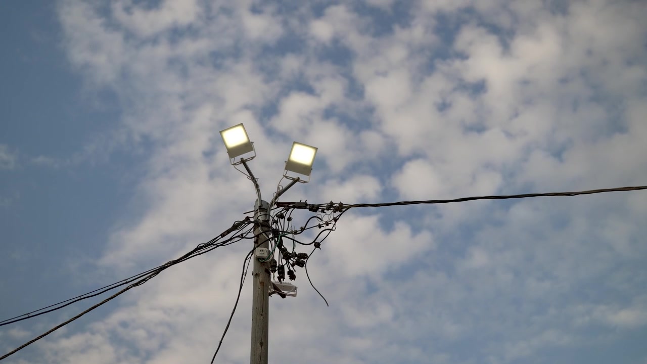 Sebuah tiang lampu yang menyala dengan kabel listrik kusut berdiri di latar belakang langit berawan sebagian