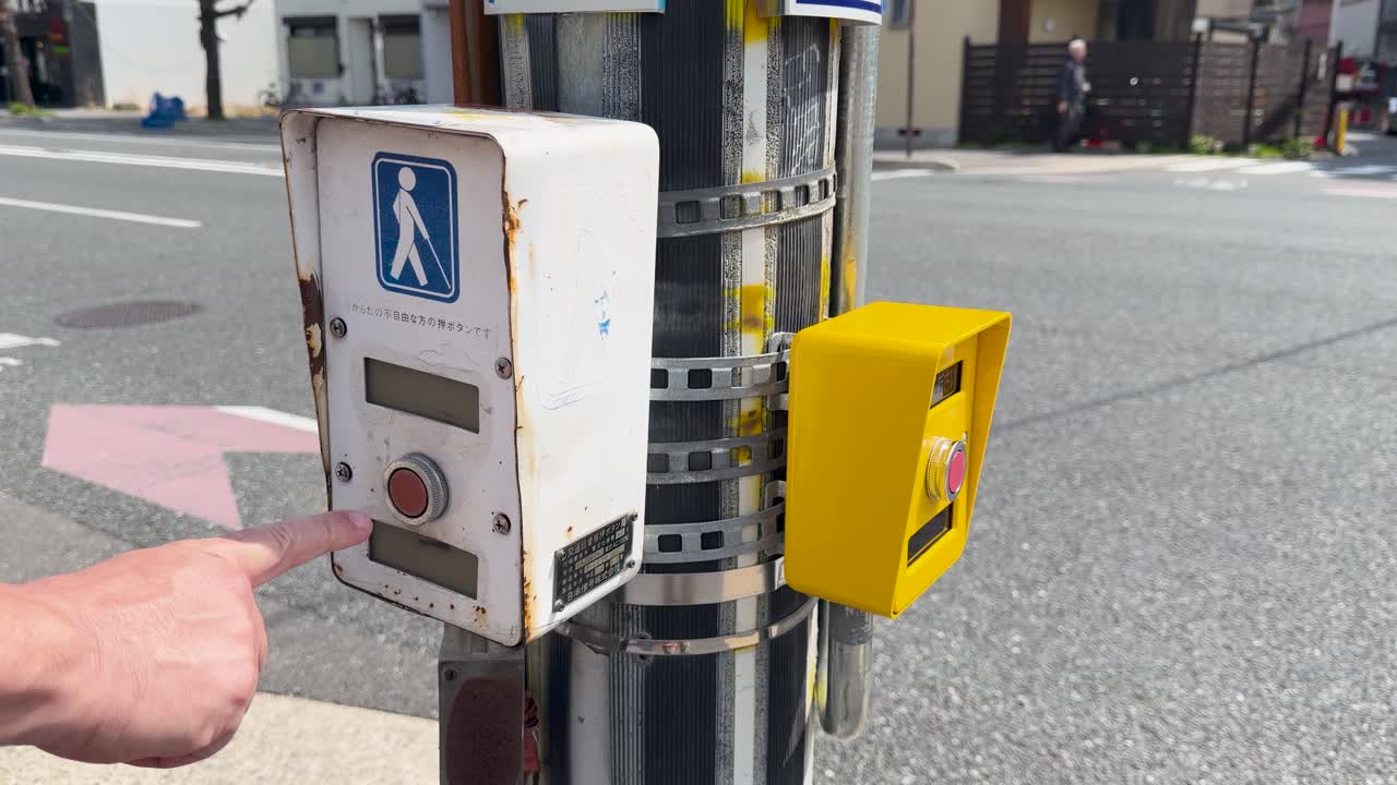 Primo piano di una mano che preme un pulsante del semaforo pedonale su una strada giapponese soleggiata