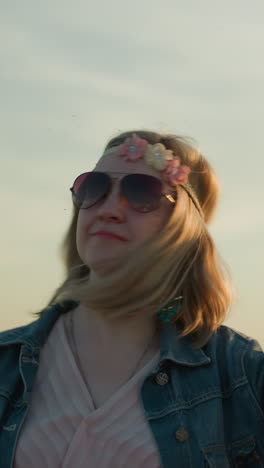 una mujer alegre con una cinta floral en la cabeza y gafas de sol se ve bailando en un campo abierto