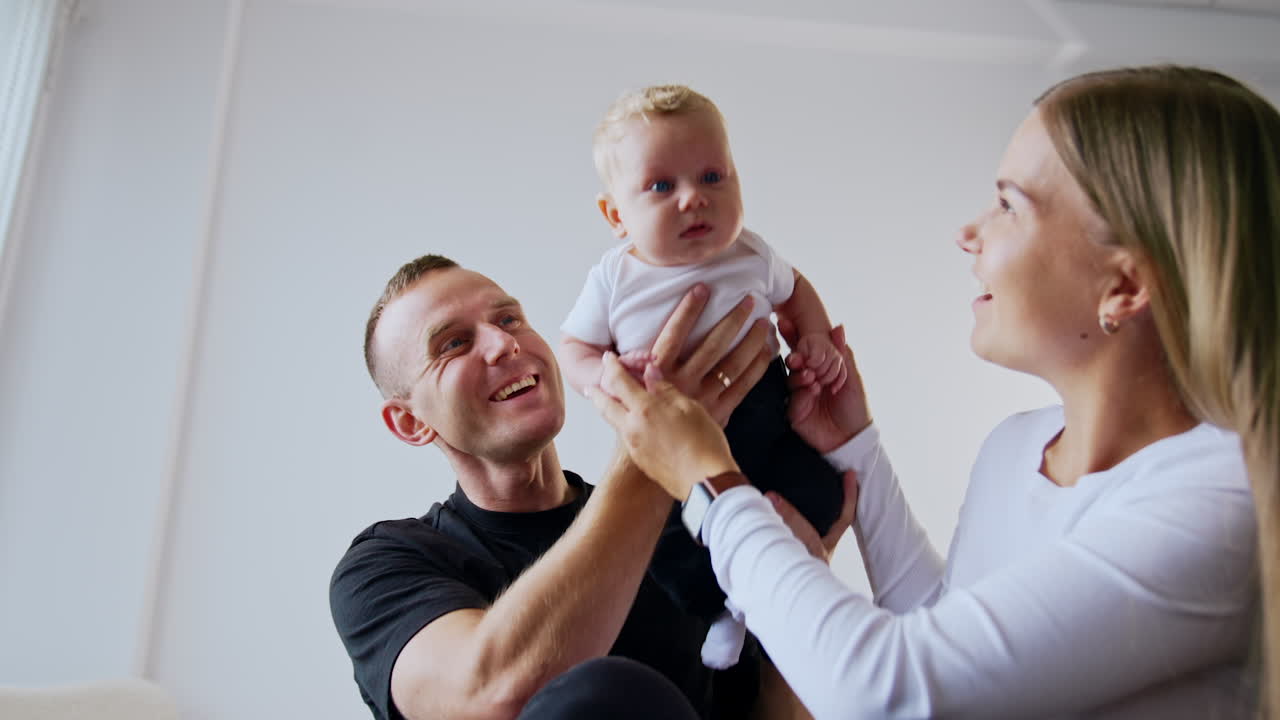 Smiling Caucasian dad kisses his adorable blond infant son. Loving parents lift their beautiful baby up. Low angle view.