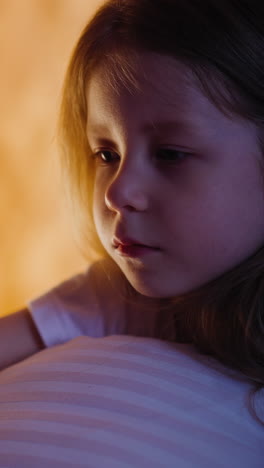 sad little girl with dimly lit half-face hugging pillow cries suffering from loneliness on bed near illuminated by play house orange light in semidark room slow motion