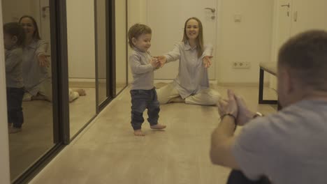 mother and her baby at one end of the corridor at home while the father encouraging the baby to walk to the other end.