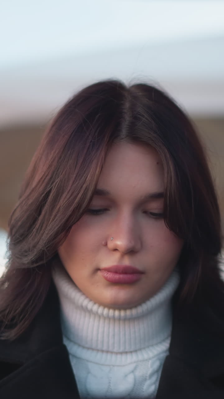 mujer joven sentada en un entorno al aire libre con el viento balanceando suavemente su cabello, profundamente en el pensamiento, ella está vestida con un cuello de tortuga blanco y abrigo negro con fondo borroso