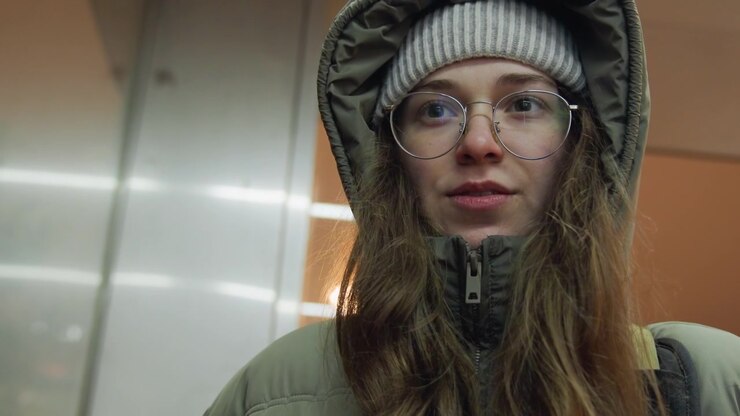 Smiling white woman in beanie closeup, candid conversational shot in subway with glasses and nose piercing, warm evening lights, relaxed winter coat and hood, leaning toward camera with approachable