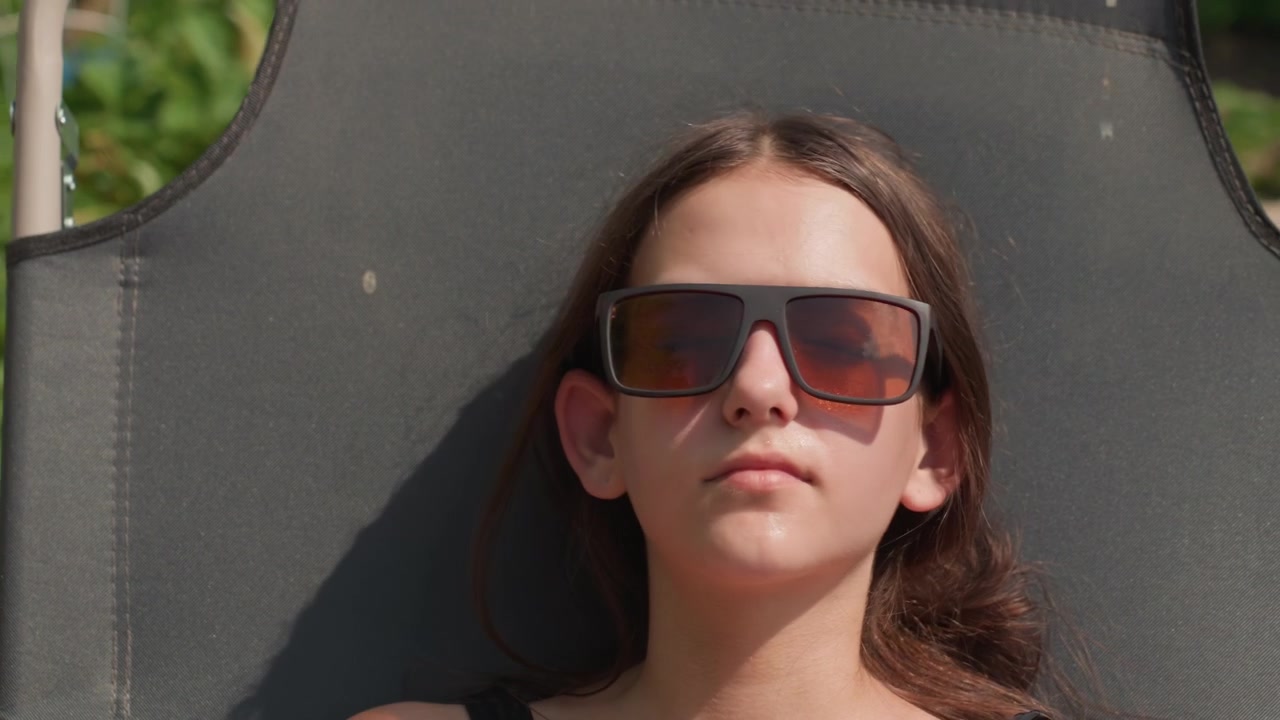 Young woman relaxes on black outdoor chair wearing sunglasses and casual clothing. scene is set in bright daylight with greenery visible outdoors capturing her calm expression as woman rests