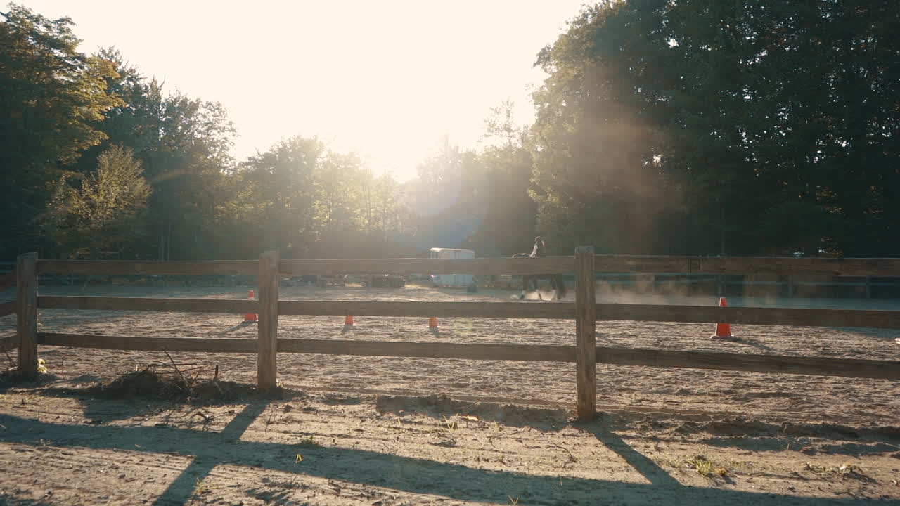 A horse and rider ride slowly in the background of a dusty arena during sunset with a fence in front.