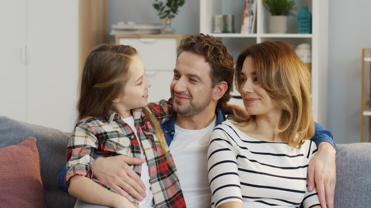 Portrait Of The Happy Good Looking Young Mother And Father Sitting On The Sofa At Home With Their Teenage Pretty Daughter, Smiling And Posing To The Camera