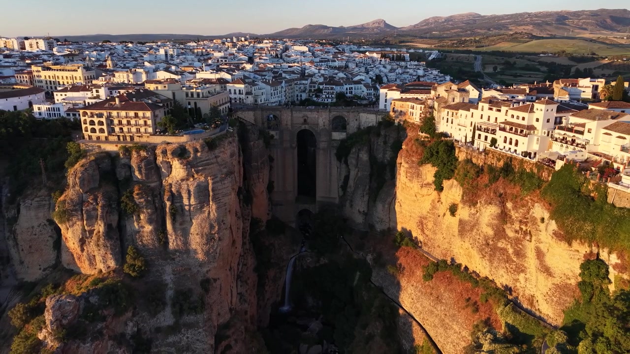 Drone aerial view of Puente Nuevo Bridge crossing El Tajo Gorge in Ronda, Spain during golden hour with dramatic cliffs, waterfall, and historic old town