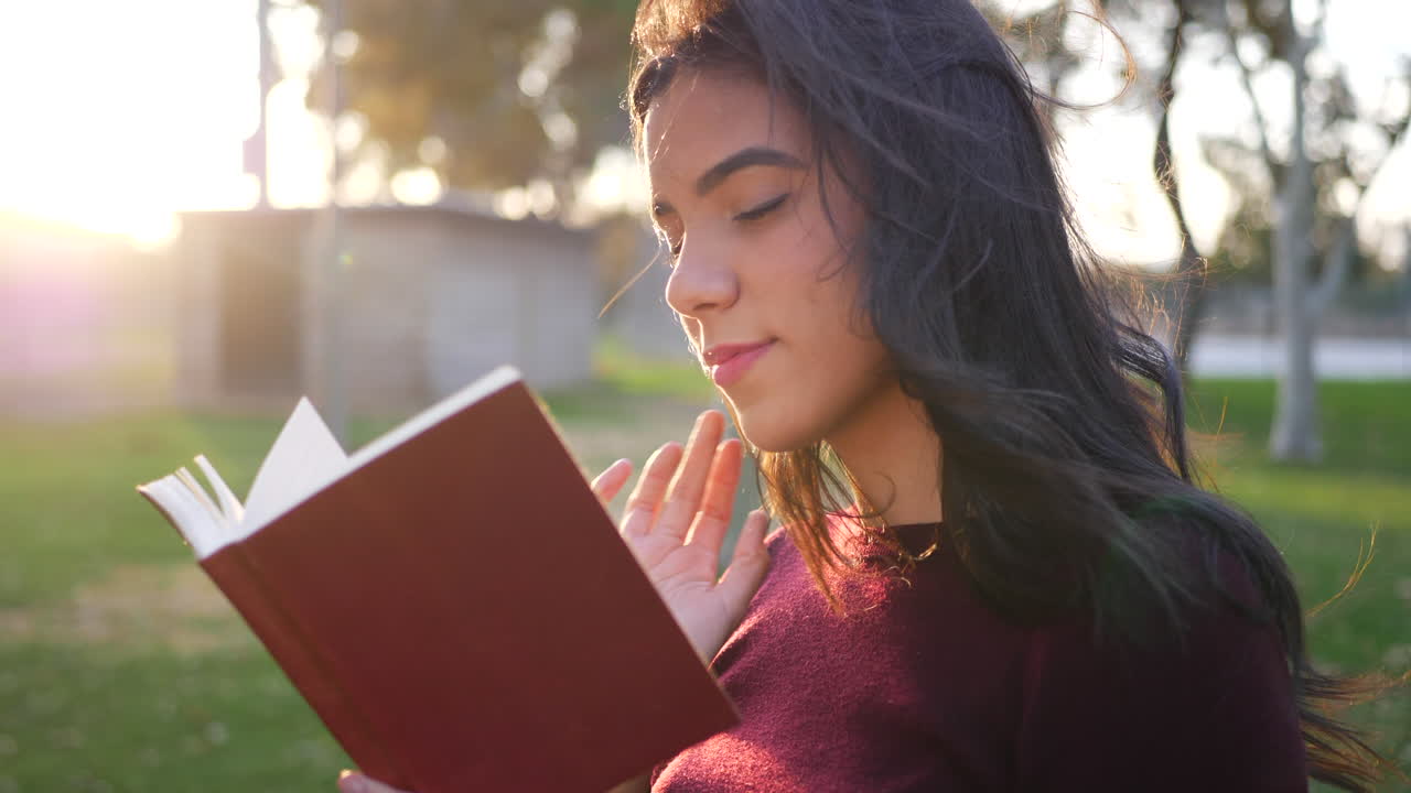 A young woman college student reading a story book outdoors in the park at sunset SLOW MOTION