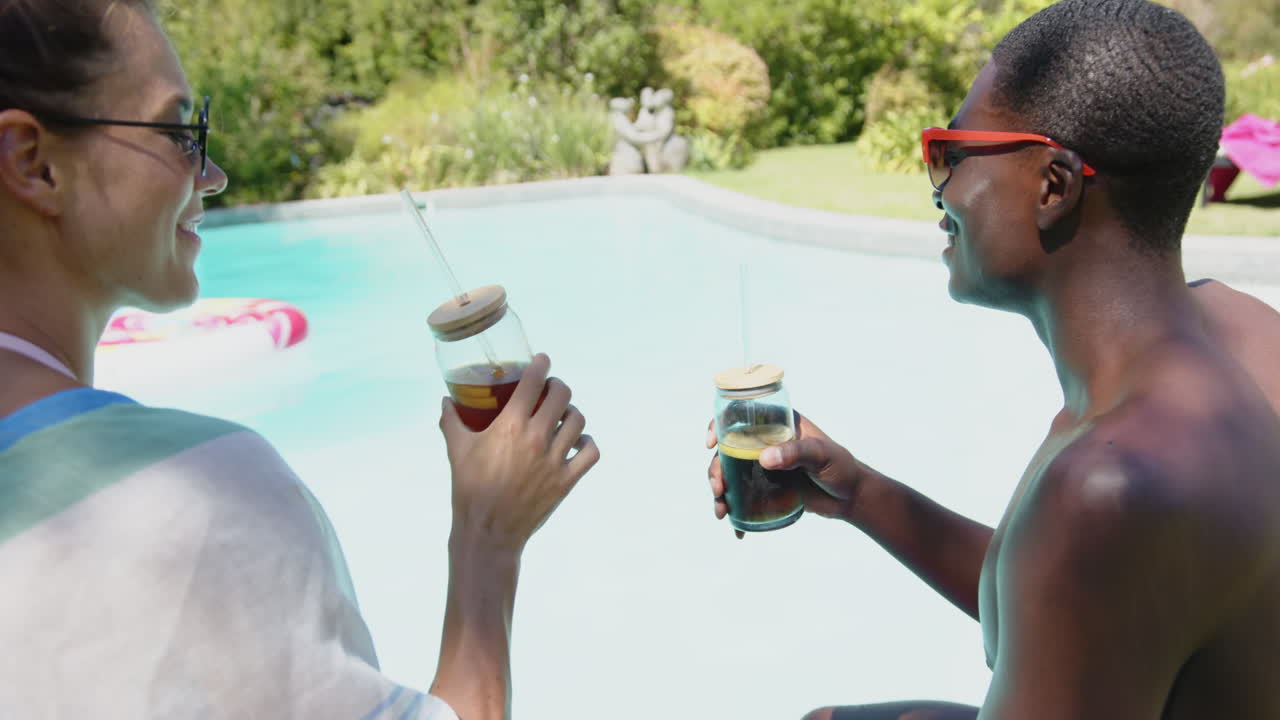 Enjoying refreshing drinks, diverse couple relaxing by pool on sunny day