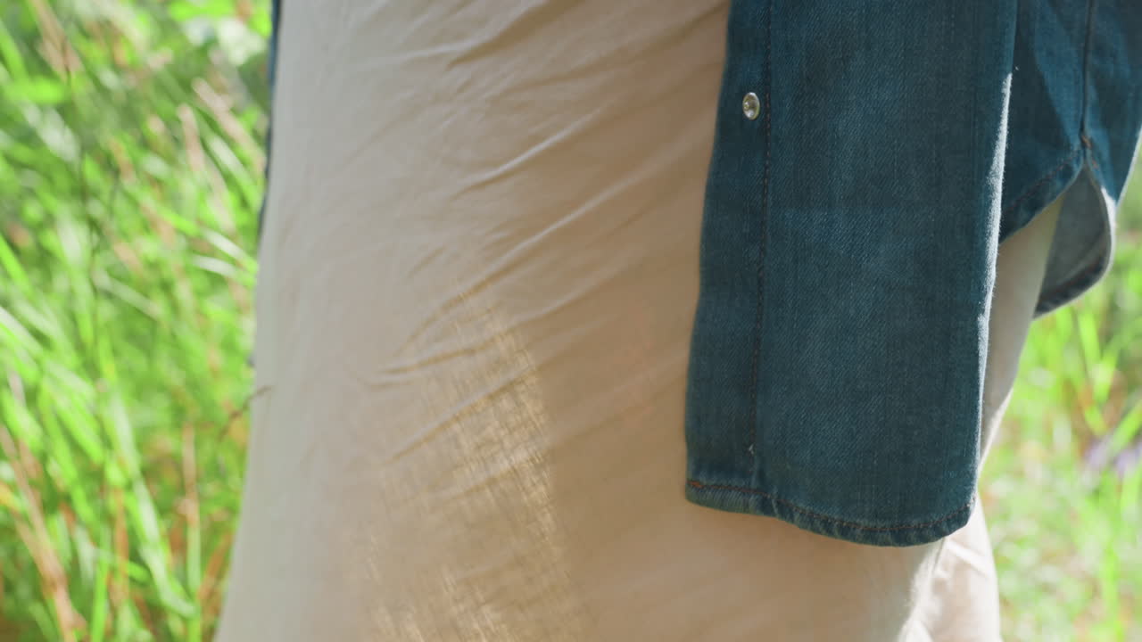 Lower view of lady wearing flowing gown and blue jacket standing outdoors under warm sunlight, soft breeze moving through tall green grass, capturing peaceful natural atmosphere