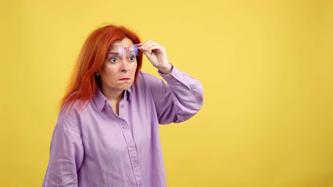 A red-haired woman with a confused expression struggles to see while holding her glasses