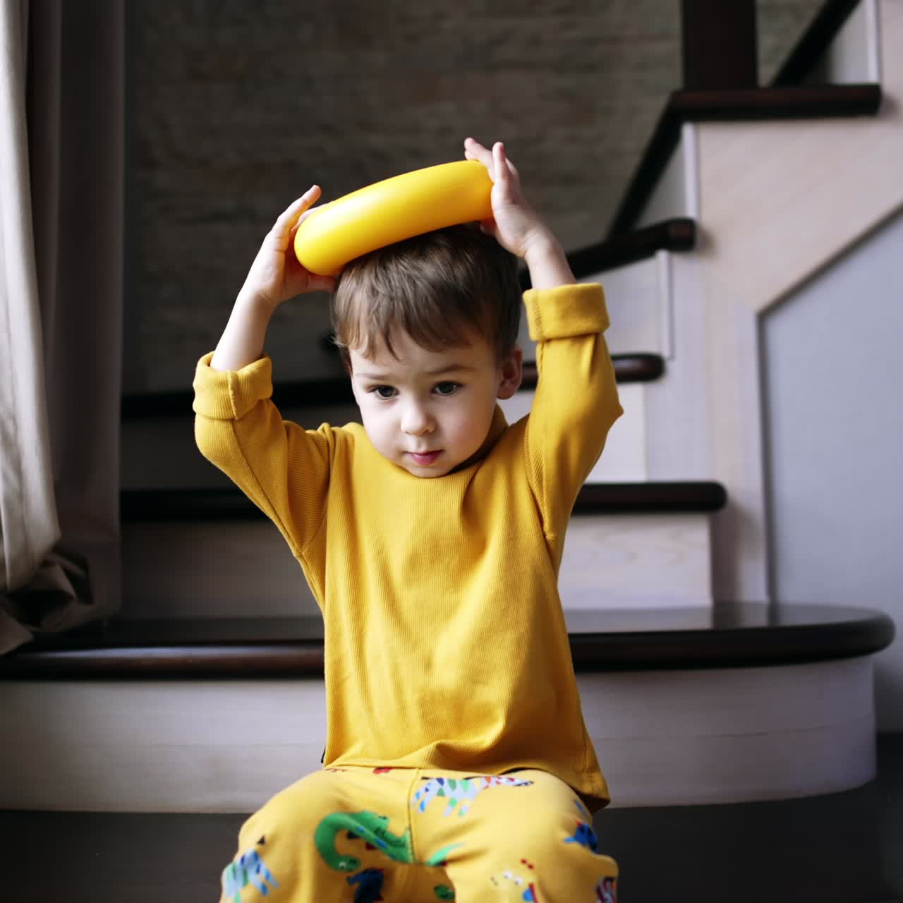 Funny toddler boy sitting on the stairs holding the ring from pyramid on his head. Kid playing at home with toys