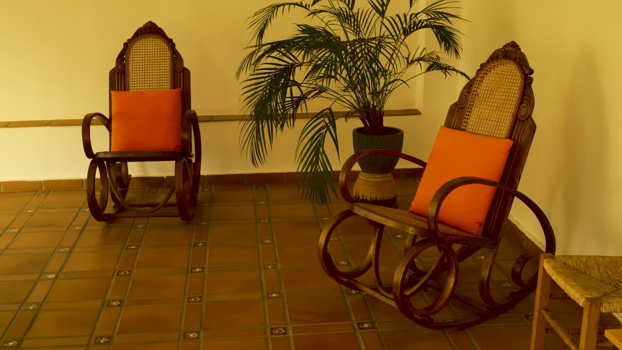 Two wooden rocking chairs with orange cushions sit beside a potted plant on a tiled floor