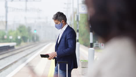 business commuters on railway platform with mobile phones wearing ppe face masks during pandemic