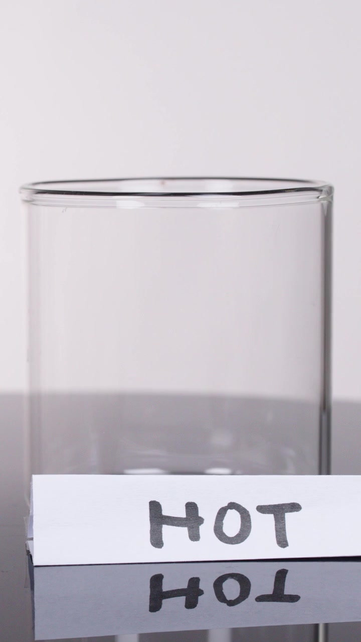 Una mano posiziona un foglio etichettato "HOT" accanto a un becher trasparente su una superficie riflettente, sotto una luminosa illuminazione da studio.