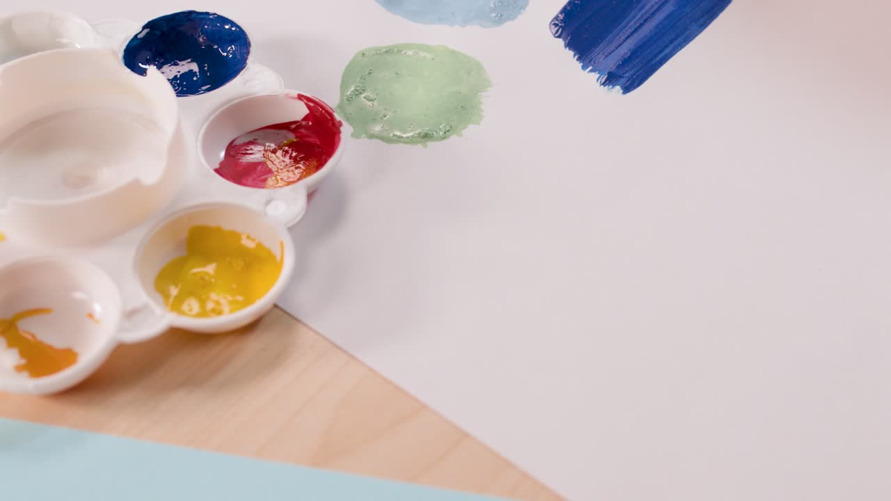 Child uses paintbrush to create colorful shapes on paper, bright lighting, overhead camera angle