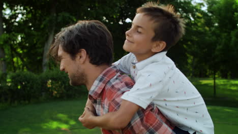 Father-spinning-around-in-park-with-son-on-back.-Funny-family-having-fun-outside